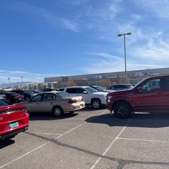 The Pit - View from parking lot - A parking lot with multiple vehicles, including a red sedan, a beige sedan, and a red pickup truck, is shown in the foreground. The lot is adjacent to a large, modern building with a flat roof, and mountains are visible in the distance. The sky is mostly clear with some wispy clouds, and tall light poles are present throughout the parking area. - Stadiums & Arenas Near Me - Albuquerque, New Mexico