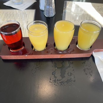 Evie’s - Mimosa flight - Four drinks are presented on a wooden flight paddle, with one red and three yellow beverages, on a dark table in a restaurant setting. - Beer, Wine & Spirits Near Me - Westerly, Rhode Island