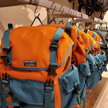 L.L.Bean - Several orange and blue L.L.Bean backpacks are arranged on a metal store rack, each hanging from a metal hook. The backpacks feature multiple pockets, adjustable straps, and buckles. In the background, other backpacks in different colors are displayed on shelves, indicating a retail environment focused on outdoor gear and accessories. - Outdoor Gear Near Me - Cincinnati, Ohio