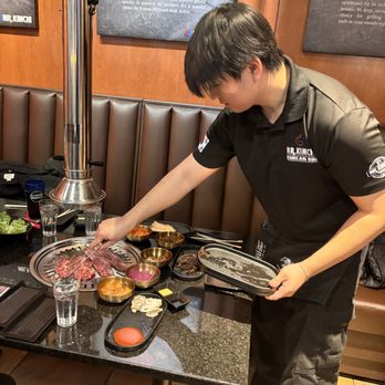 Mr Kimchi - A staff member at Mr. Kimchi Korean BBQ is placing raw meat on a grill at a restaurant table, surrounded by various side dishes and condiments. - Barbeque Near Me - Algonquin, Illinois