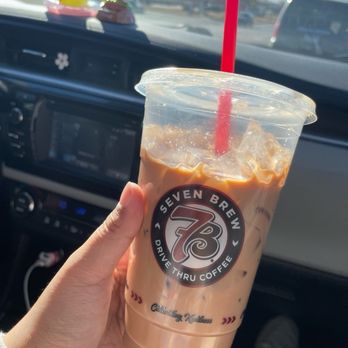 7 Brew Coffee - A person is holding a large plastic cup of iced coffee from Seven Brew Drive Thru Coffee, featuring the company's logo on the cup. The drink has a red straw and is partially consumed. The background shows the interior of a vehicle, including the dashboard and windshield, with a view of the road and traffic signals outside. Two small decorative figures are placed on the dashboard. - Coffee & Tea Near Me - Champaign, Illinois