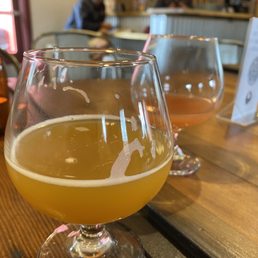 Two glasses of beer are positioned on a wooden table inside a brewery or bar. The glass in the foreground contains a hazy yellow beer, while the background glass holds an amber-colored beer. The setting includes indoor seating with chairs and a bar area visible in the background, where two people are seated and engaged in activities. The environment appears casual and relaxed, with a menu or sign on the table.