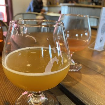 Knotted Root Brewing - Two glasses of beer are positioned on a wooden table inside a brewery or bar. The glass in the foreground contains a hazy yellow beer, while the background glass holds an amber-colored beer. The setting includes indoor seating with chairs and a bar area visible in the background, where two people are seated and engaged in activities. The environment appears casual and relaxed, with a menu or sign on the table. - Breweries Near Me - Nederland, Colorado