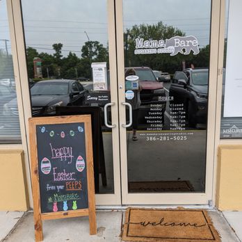 Mama Bare Sugaring Studio - Ormond Beach - The image shows the entrance of Mama Bare Sugaring Studio, featuring glass doors with business hours and a sign. A chalkboard sign with Easter greetings and decorations is placed outside. - Sugaring Near Me - Ormond Beach, Florida