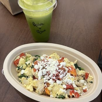 t.Loft - Alive & Alert Sunrise Bowl - A salad with scrambled eggs, chopped tomatoes, and greens, topped with crumbled cheese, served in a disposable bowl. A green beverage in a clear cup with a lid is placed behind it. - Juice Bars & Smoothies Near Me - Lawrence, Kansas