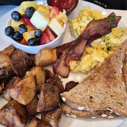 All American Breakfast - A breakfast plate featuring scrambled eggs, crispy bacon, roasted potatoes, toasted bread, and a side of mixed fruit with syrup.
