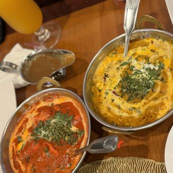 Curry Masala - Spring - Chicken Tikka Masala and Butter Chicken - The image shows two stainless steel bowls of Indian curry dishes on a wooden table. One bowl contains butter chicken with a creamy orange sauce, and the other has chicken tikka masala with a yellowish, spiced gravy, both garnished with chopped cilantro. In the background, there are drinks, including a glass of orange-colored beverage and a glass of water with ice. The setting appears to be a restaurant with a warm, wooden interior. - Indian Near Me - Spring, Texas