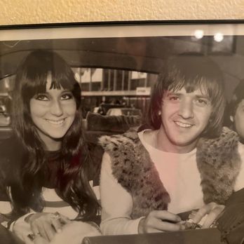 Lighthouse Tavern - A black-and-white photograph shows a woman and a man sitting side by side, smiling at the camera, with a background of a parking lot. - Bars Near Me - Chicago, Illinois