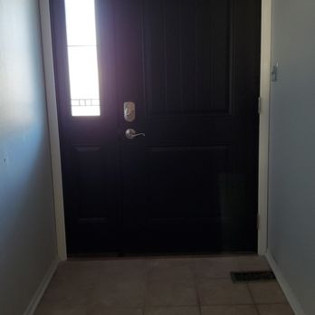 Rusco Windows & Doors - Inside front door - A black front door with a narrow, vertical window panel on the left side, featuring a decorative grid pattern. The door is framed by a white border and is situated in an entryway with tiled flooring and light-colored walls. A door handle and lock are visible on the right side of the door, and a floor vent is located on the lower right corner of the image. - Windows Installation Near Me - Downers Grove, Illinois