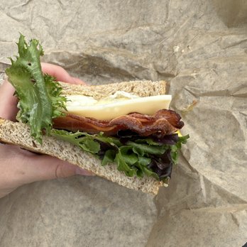 Common Ground - Hampden special on sourdough - A sandwich featuring lettuce, cheese, bacon, and bread is held in a person's hand, with a background of crumpled brown paper. - Coffee & Tea Near Me - Baltimore, Maryland