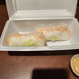 Thai sliding rolls - Two shrimp spring rolls with lettuce are placed in a white foam takeout container, accompanied by a small cup of dipping sauce.