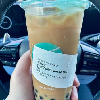 T4 - Bruh this is not Almond Milk Tea ($6.55 for a Large with Pearls) - A large plastic cup containing almond milk tea with tapioca pearls is held in front of a car's steering wheel. The drink has a light brown color, and the pearls are visible at the bottom. The cup has a plastic lid with a straw inserted, and a paper sleeve with the order details. The background shows the interior of a vehicle, including the steering wheel and dashboard. - Bubble Tea Near Me - Millbrae, California