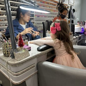 V Nails - A young girl with long, wavy hair and a pink bow in her hair is seated at a nail salon station, receiving a manicure. The station is decorated with small Christmas trees and glittery containers, and a bright lamp illuminates the area. Behind her, a technician with a face mask and glasses is working, with a wall of nail polish bottles visible in the background. The setting appears to be a busy nail salon with multiple workstations. - Nail Salons Near Me - Avon, Indiana