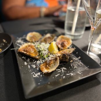 Flavorture - Charbroiled Oysters - A black square plate holds several charbroiled oysters topped with seasoning, garnished with shredded cheese, microgreens, and lemon wedges, with drinks and a person in the background. - Bars Near Me - Washington, DC, District of Columbia