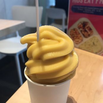 RASA - Masala Chai mango lassi swirl soft serve - A cup of soft serve ice cream with a mango lassi flavor, characterized by a yellow color and a swirl shape, is positioned on a light wooden table. The background includes a menu and some chairs, indicating a casual restaurant or cafe environment. The ice cream is served in a plain white cup, and a small stick is inserted into the swirl for support. - Indian Near Me - Fairfax, Virginia