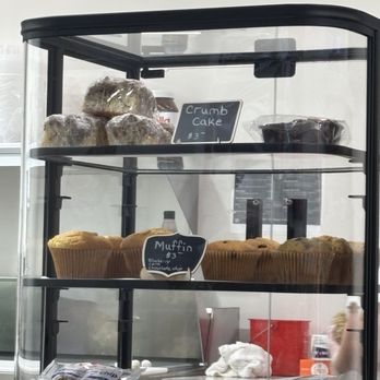 Time 4 Bagels - Nonbagel eaters can have a pastry if you'd like! - A clear glass bakery display case showcases baked goods such as crumb cake on the top shelf and muffins on the middle shelf. The crumb cake is labeled with a small sign indicating a price of $3, and the muffins are labeled as blueberry and chocolate chip, also priced at $3. The case is situated on a countertop with some packaging and a cloth visible at the bottom. The background includes a white wall and some kitchen items. - Bagels Near Me - Gainesville, Virginia