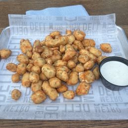 A tray filled with fried chicken nuggets is placed on a wooden table. The nuggets are golden-brown and appear crispy, with some showing herbs or seasoning. A small black cup containing a white, creamy dipping sauce is positioned on the right side of the tray. The tray is lined with branded paper featuring the word 'DEFT' repeatedly. The background includes a metal surface and some equipment, indicating an indoor or outdoor dining setting.