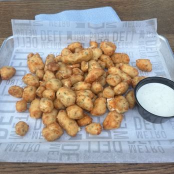 Pilot Project Brewing - Milwaukee - A tray filled with fried chicken nuggets is placed on a wooden table. The nuggets are golden-brown and appear crispy, with some showing herbs or seasoning. A small black cup containing a white, creamy dipping sauce is positioned on the right side of the tray. The tray is lined with branded paper featuring the word 'DEFT' repeatedly. The background includes a metal surface and some equipment, indicating an indoor or outdoor dining setting. - Brewpubs Near Me - Milwaukee, Wisconsin