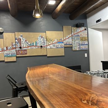 El Vado Taproom - Nice cozy bar - The image shows a bar interior with a polished, natural wood bar counter in the foreground. The background includes a dark gray wall decorated with license plates and license plate art arranged in a creative display. The ceiling has large wooden beams and modern pendant lighting. The setting appears to be a casual, rustic-style bar with a focus on unique decor elements. - Beer Bar Near Me - Albuquerque, New Mexico