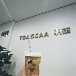 Brown Sugar Bubble Milk - A hand is holding a plastic cup of brown sugar bubble milk with a straw, positioned in front of a white wall. The wall displays the Tsaocaa logo and Chinese characters, indicating the location is a Tsaocaa bubble tea shop. The drink appears to be a milk tea with dark tapioca pearls at the bottom, and the cup has a Tsaocaa logo printed on it.