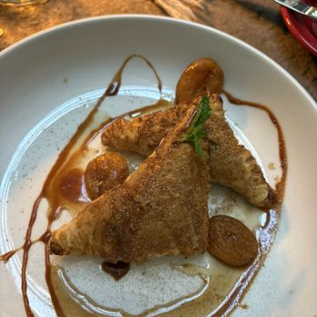 Amaya - Desserts - Three fried samosas are arranged on a white plate, garnished with a small mint leaf. The samosas are golden brown and crispy, with a drizzle of sauce around them. Sliced fruit pieces are placed on the plate, and the dish is presented on a table with a colorful cloth and utensils nearby. - New American Near Me - Santa Fe, New Mexico