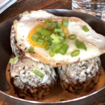 Empire - Empire Tots - A food presentation showing a fried egg with a runny yolk, garnished with chopped green onions, placed on top of two rice balls coated with a creamy sauce. The dish is served in a metal bowl on a wooden table, with a glass of water and plates visible in the background. - Burgers Near Me - Naperville, Illinois
