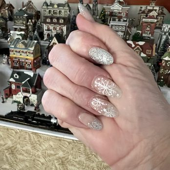 LA Nails - A hand with glittery and snowflake-themed nail art is held in front of a detailed miniature Christmas village display. - Nail Salons Near Me - Yarmouth, Maine
