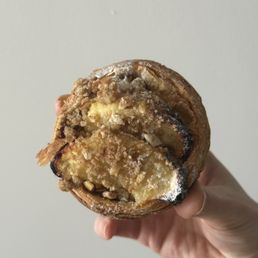 Apple pastry - A pastry featuring a crumbly topping and apple filling is held in a person's hand. The pastry appears baked with a golden-brown crust and visible apple pieces inside. The background is plain and neutral, emphasizing the pastry's texture and appearance.
