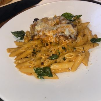 Yard House - Fontina Penne - A dish of penne pasta is presented on a white plate, topped with melted cheese and a tomato-based sauce. Fresh spinach leaves are scattered around the pasta, and the dish appears to be served in a restaurant setting with a wooden table and other food items visible in the background. - New American Near Me - Washington, DC, District of Columbia