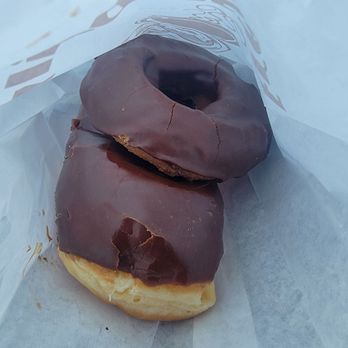 Donut Tree - Inside the bag - The image shows two donuts with chocolate glaze inside a paper bag. One donut is slightly larger and has a cracked chocolate coating, while the other appears smaller with a smoother surface. The donuts are resting on a piece of white paper or parchment inside the bag, which has some printed text partially visible. The background is plain and light-colored. - Donuts Near Me - Oroville, California