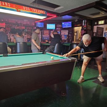 R Place - A man is playing pool in a bar with a USC Trojans sign overhead, while other patrons sit at the bar in the background. - Dive Bars Near Me - Pasadena, California