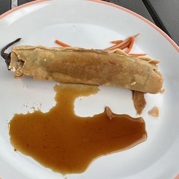 Pesca Vilano - Lumpia - A fried spring roll is placed on a white plate with a pink rim, accompanied by a pool of soy or dipping sauce. Some shredded carrots are visible on the plate, and the plate is set on a dark outdoor surface, possibly a table or bench. - Seafood Near Me - St. Augustine, Florida