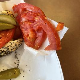 Mushy tomatoes - The image shows slices of fresh tomato, pickles, and a poppy seed bagel on a white paper-lined tray. The tray is placed on a brown surface, and the food items are arranged with the tomato slices and pickles visible. The setting appears to be a casual dining environment, consistent with Herm's Palace diner in Skokie, IL.