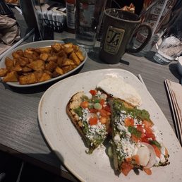 Avocado, toast, and Crispy Seasoned Home Fries - A breakfast plate features avocado toast with diced tomatoes, green onions, and a fried egg, served on a white plate. A bowl of seasoned home fries is nearby, with drinks including water and coffee on the table.
