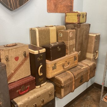 Buffalo Exchange - I love it! - A group of vintage suitcases of different sizes, colors, and styles are arranged on a shelf and against a wall in an indoor setting. The suitcases exhibit signs of age, including scuffs, scratches, and faded fabric. The collection includes hard-sided and soft-sided cases, some with handles and metal clasps, arranged in a somewhat haphazard manner. - Women's Clothing Near Me - Austin, Texas