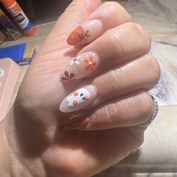 Fall Oct set - Close-up of a hand displaying decorated nails with intricate nail art. The nails feature floral designs, a butterfly, and metallic accents in orange, white, and brown. The background shows a workspace with nail supplies and tools, indicating a manicure session.