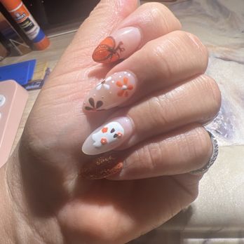 Tiny Nails - Fall Oct set - Close-up of a hand displaying decorated nails with intricate nail art. The nails feature floral designs, a butterfly, and metallic accents in orange, white, and brown. The background shows a workspace with nail supplies and tools, indicating a manicure session. - Nail Salons Near Me - Chicago, Illinois