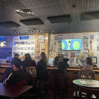 The Quarry Thornton - Inside bar - The image shows the interior of a bar with a dark ceiling and walls decorated with black-and-white photos, framed pictures, and guitars hanging on the wall. There are multiple patrons seated at the bar and tables, some engaged in conversation and others looking at their phones. A large TV screen is mounted on the wall, displaying a blue and green graphic. The bar area has a chalkboard sign advertising open mic and Thursday specials, and the lighting includes hanging pendant lights. - Bars Near Me - Thornton, Illinois