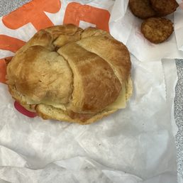 Maple sugar bacon sandwich with a side of hash browns - A croissant sandwich with melted cheese inside is shown on Dunkin' Donuts branded paper, accompanied by three hash browns on a separate piece of paper. The items are placed on a gray, speckled countertop.