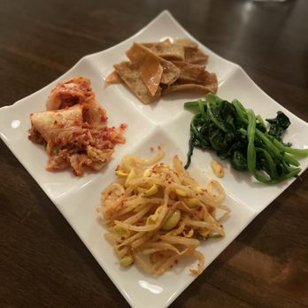 The Lotus - The free appetizer - A white, four-compartment plate contains Korean dishes including kimchi, seasoned bean sprouts, cooked spinach, and sliced beef. The plate is placed on a dark wooden table, with a glass of water and other tableware visible in the background. The dishes are arranged separately, showcasing traditional Korean side dishes. - Korean Near Me - Ithaca, New York