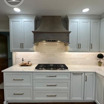 Cabinet Tree Studio - A kitchen with white cabinetry and gold handles, a gray range hood centered above a black gas cooktop, and a beige brick-style backsplash. The countertop is white with a small wooden tray and a potted plant as decor. The ceiling has recessed lighting, and the floor is dark wood. - Interior Design Near Me - Plainfield, Illinois
