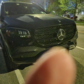 OG Garage - After - The image shows a black Mercedes-Benz SUV parked on a street during nighttime. The vehicle's front grille and headlights are visible, with the Mercedes-Benz emblem prominently displayed. The background includes trees with green leaves, a streetlamp illuminating the scene, and residential houses. A finger is partially covering the lower part of the camera lens, obscuring part of the vehicle's front bumper and license plate. - Auto Customization Near Me - Brooklyn, New York