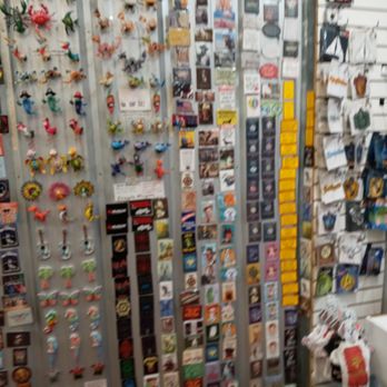 Magnet Force - The image shows a retail display with multiple vertical panels filled with small souvenirs, magnets, and decorative items. The items include colorful figurines, keychains, and various small collectibles. The display is densely packed, with some items hanging on hooks and others attached to the panels. Additional items and packaging are visible on the right side, including postcards and small packaged products. The setting appears to be inside a gift or souvenir shop, likely in a tourist area. - Gift Shops Near Me - Saint Augustine, Florida