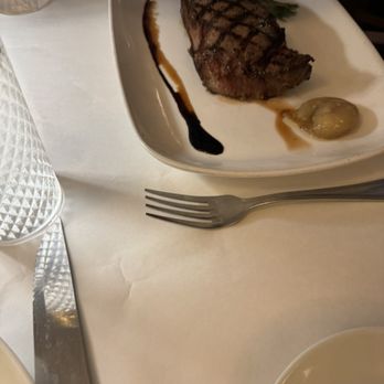 Fauna - Steak - A square white plate contains a grilled steak with visible grill marks, accompanied by a small onion and a sauce drizzle. The plate is placed on a white tablecloth, with a fork positioned nearby and a textured glass of water partially visible on the left. The setting suggests a dining environment with a focus on the presentation of the steak dish. - New American Near Me - Westhampton Beach, New York