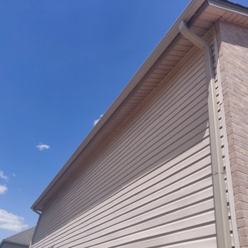 Rain Tech Gutter Solutions - New Gutters - A building's exterior corner is shown, featuring beige vinyl siding on one side and a brick wall on the adjacent side. The roof has a gutter system with downspouts, and the sky in the background is clear and blue with a few small clouds. The image emphasizes the construction and materials used in the building's exterior. - Gutter Services Near Me - Liberty hill, Texas