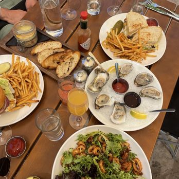 Agustin Kitchen - A dining table features oysters on a bed of ice, salads, sandwiches with French fries, bread, and various beverages, including water and cocktails. - New American Near Me - Tucson, Arizona