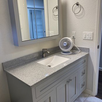EZ Baths - Tons of colors/finishes to choose from- clearly I love gray - A bathroom vanity features a granite countertop with an integrated sink, a faucet, and a mirrored cabinet above. A small, portable fan is placed on the countertop near the wall, with its cord plugged into an electrical outlet. The wall has a towel ring and a double light switch. The mirror cabinet reflects a doorway and part of the bathroom interior. - General Contractors Near Me - Hayward, California