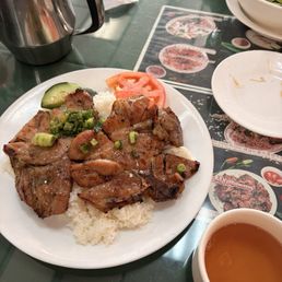 A white plate contains grilled meat served over a bed of white rice, garnished with chopped green onions. Slices of tomato and cucumber are placed on the side of the plate. The setting includes a green table with a menu, a bowl of salad, a cup of tea, and a metal teapot. The background shows a tissue and part of the restaurant interior.