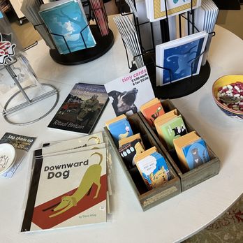 Double Dog Studios - A round table features a variety of items including books titled 'Downward Dog' and 'Ben Edge and The Museum of British Folklore,' along with postcards and a bowl of assorted candies with a spoon. A card display rack holds greeting cards with animal illustrations, and a small dish contains a few more items. The setting appears to be a retail or gift shop with organized displays and a cozy atmosphere. - Art Galleries Near Me - Carnegie, Pennsylvania