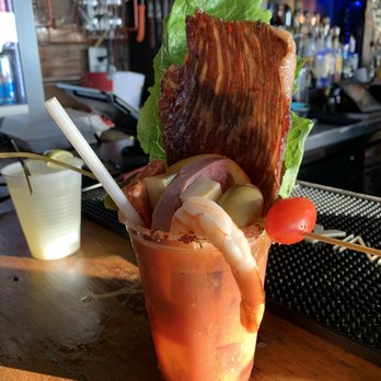 Low Key Hideaway - Loaded Bloody Mary - A Bloody Mary cocktail is presented in a tall glass with a variety of garnishes such as a strip of bacon, a cooked shrimp, lettuce, pickles, and a cherry tomato on a skewer. The drink is placed on a wooden bar counter with a straw, and the background shows bar equipment and a dimly lit bar area. - Bars Near Me - Cedar Key, Florida